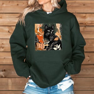 Puede incluir: Sudadera con capucha verde oscuro con una ilustraci&oacute;n de gato negro y elementos oto&ntilde;ales. El dise&ntilde;o incluye una calabaza y hojas en tonos naranja y crema. La sudadera tiene un bolsillo delantero y una capucha.