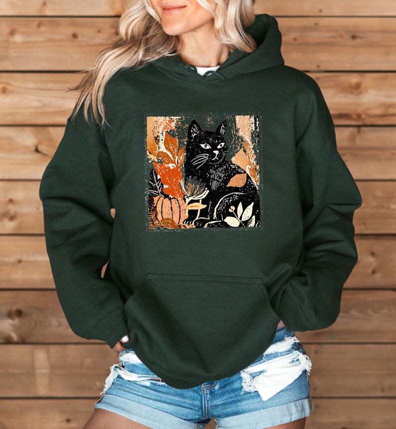 Puede incluir: Sudadera con capucha verde oscuro con una ilustraci&oacute;n de gato negro y elementos oto&ntilde;ales. El dise&ntilde;o incluye una calabaza y hojas en tonos naranja y crema. La sudadera tiene un bolsillo delantero y una capucha.