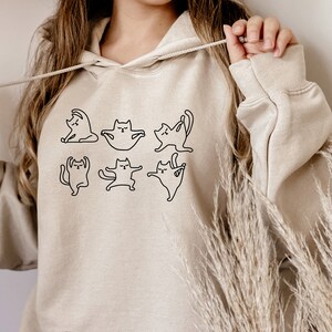 May include: A beige hoodie with a black outline of cats doing yoga poses. The hoodie has a drawstring hood.