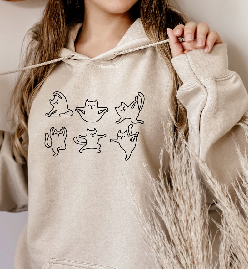 May include: A beige hoodie with a black outline of cats doing yoga poses. The hoodie has a drawstring hood.
