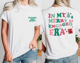 Op de afbeelding: Wit T-shirt met de tekst "FUTURE MRS SCOTT" in groen op de voorkant. De achterkant van het shirt heeft de tekst "IN MY MERRY & ENGAGED ERA" in roze, groen en rood, met kerstthema graphics.
