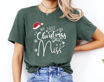 Op de afbeelding: Donkergroen t-shirt met de witte tekst "My Last Christmas as a Miss". Het ontwerp bevat een kerstmuts, sneeuwvlokken en een ring. Het shirt heeft een ronde hals.