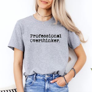 May include: A light gray t-shirt with the text "Professional Overthinker" printed in black. The shirt is a crew neck style and is made of a soft material. The model is wearing blue jeans.