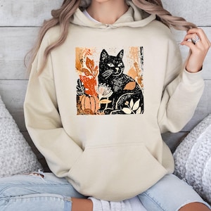 Puede incluir: Sudadera con capucha beige con un dise&ntilde;o de gato negro rodeado de hojas de oto&ntilde;o y una calabaza. El dise&ntilde;o utiliza tonos naranja, marr&oacute;n y blanco. La sudadera tiene un bolsillo de canguro y una capucha con cord&oacute;n.