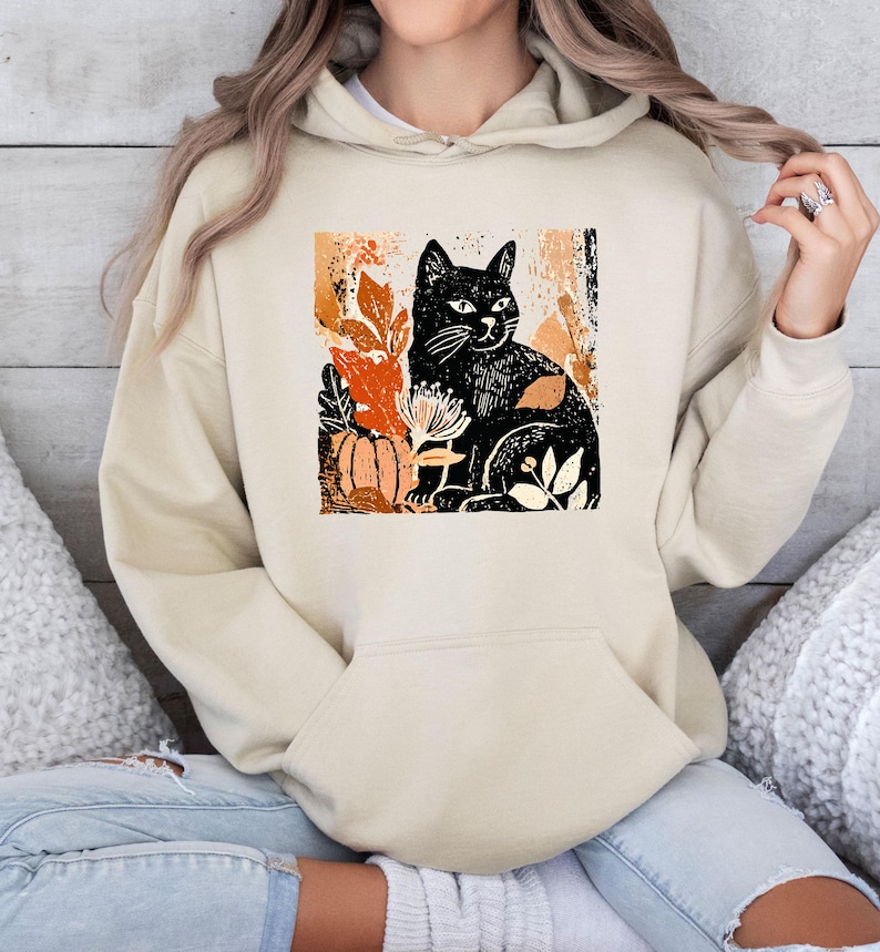 Puede incluir: Sudadera con capucha beige con un dise&ntilde;o de gato negro rodeado de hojas de oto&ntilde;o y una calabaza. El dise&ntilde;o utiliza tonos naranja, marr&oacute;n y blanco. La sudadera tiene un bolsillo de canguro y una capucha con cord&oacute;n.