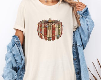 Puede incluir: Camiseta color crema con un diseño de adorno navideño hecho de libros. El adorno está envuelto con luces de cadena. Los libros son rojos, verdes y marrones con las palabras "Merry Christmas" impresas en algunos.