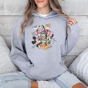 May include: A light gray hooded sweatshirt featuring a colorful graphic of Disney characters including Mickey Mouse, Minnie Mouse, Donald Duck, and Goofy. The graphic has character signatures. The sweatshirt is made of a soft material.