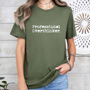May include: Olive green t-shirt with the text "Professional Overthinker" in a white, typewriter-style font. The shirt is a short-sleeved crew neck. The model is wearing blue jeans and bracelets.