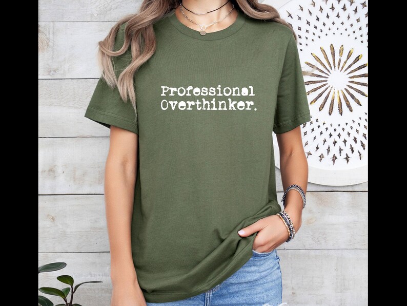 May include: Olive green t-shirt with the text "Professional Overthinker" in a white, typewriter-style font. The shirt is a short-sleeved crew neck. The model is wearing blue jeans and bracelets.