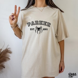 May include: A beige oversized t-shirt with black text that reads "Parker Est. 2001" and a black spider graphic.