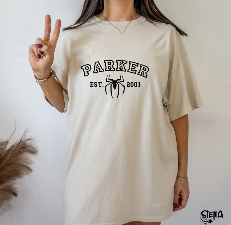May include: A beige oversized t-shirt with black text that reads "Parker Est. 2001" and a black spider graphic.