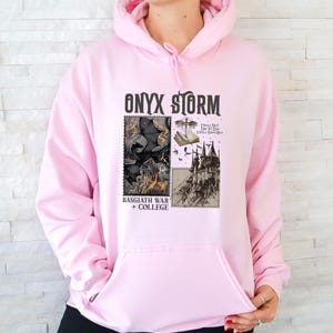Peut inclure: Sweat-shirt rose avec un imprim&eacute; graphique d'un dragon noir, d'un ch&acirc;teau et le texte "ONYX STORM", "I WILL NOT DIE TO DAY I WILL SAVE HIM", "BASSGIATH WAR + COLLEGE".