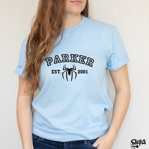 May include: Light blue t-shirt with black text that reads "Parker Est. 2001" and a black spider graphic.
