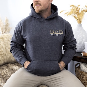 May include: Dark gray hoodie with a front pocket and a hood. The word "DAD" is printed on the chest in gold lettering, with "Oliver & Sarah" below. The hoodie is worn by a person.