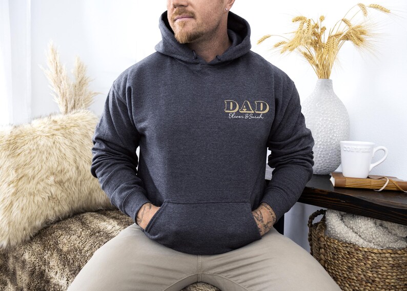 May include: Dark gray hoodie with a front pocket and a hood. The word "DAD" is printed on the chest in gold lettering, with "Oliver & Sarah" below. The hoodie is worn by a person.