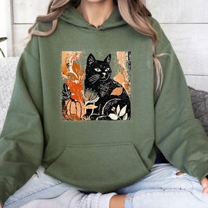 Puede incluir: Sudadera con capucha verde oliva con un dise&ntilde;o de gato negro, detalles en naranja y blanco. El gr&aacute;fico incluye una calabaza y follaje, lo que sugiere un tema oto&ntilde;al o de Halloween. La sudadera tiene un bolsillo de canguro y una capucha con cord&oacute;n.