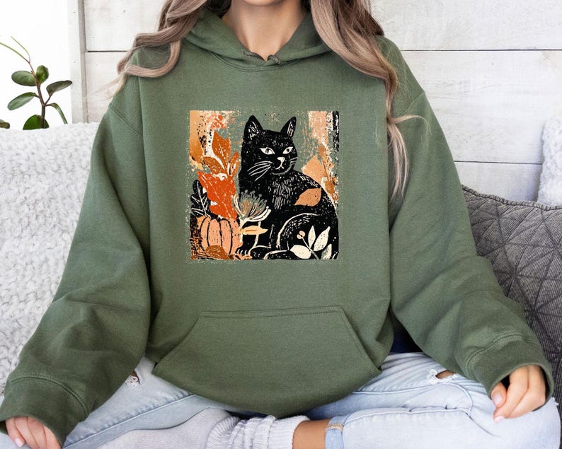 Puede incluir: Sudadera con capucha verde oliva con un dise&ntilde;o de gato negro, detalles en naranja y blanco. El gr&aacute;fico incluye una calabaza y follaje, lo que sugiere un tema oto&ntilde;al o de Halloween. La sudadera tiene un bolsillo de canguro y una capucha con cord&oacute;n.