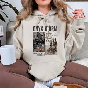 Peut inclure: Un sweat &agrave; capuche beige avec un imprim&eacute; graphique d'un ch&acirc;teau, d'un livre et d'un corbeau. Le texte "ONYX STORM" est imprim&eacute; en noir au-dessus du graphique. Le texte "BASSGIATH WAR + COLLEGE" est imprim&eacute; en dessous du graphique. Le texte "I Will Not Die For This Day I Will Save Him" est imprim&eacute; en petite police sur le c&ocirc;t&eacute; droit du graphique.