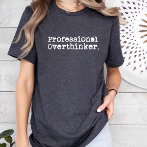 May include: Dark gray t-shirt with the text "Professional Overthinker" in white, centered on the chest. The shirt has short sleeves and a crew neck. The fabric appears soft and comfortable.