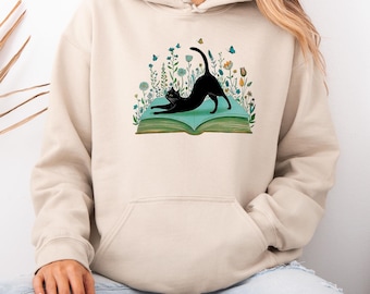 Puede incluir: Sudadera con capucha beige con un gato negro estirándose sobre un libro abierto, con ilustraciones de flores y mariposas. El diseño incluye detalles en verde, azul y amarillo. La sudadera tiene un bolsillo delantero y una capucha con cordón.