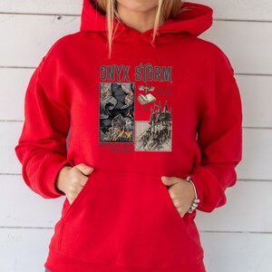 Peut inclure: Sweat-shirt &agrave; capuche rouge avec un imprim&eacute; graphique d'un ch&acirc;teau, d'un livre et d'une chauve-souris avec le texte "ONYX STORM" et "Something Wicked This Way Comes".