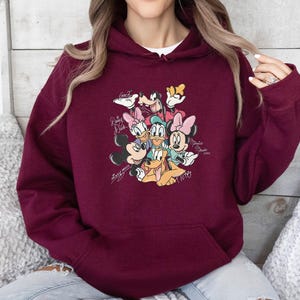 May include: Burgundy hooded sweatshirt featuring a colorful graphic of Disney characters including Mickey Mouse, Minnie Mouse, Donald Duck, Daisy Duck, Goofy, and Pluto. The design includes character signatures. A white undershirt is visible.