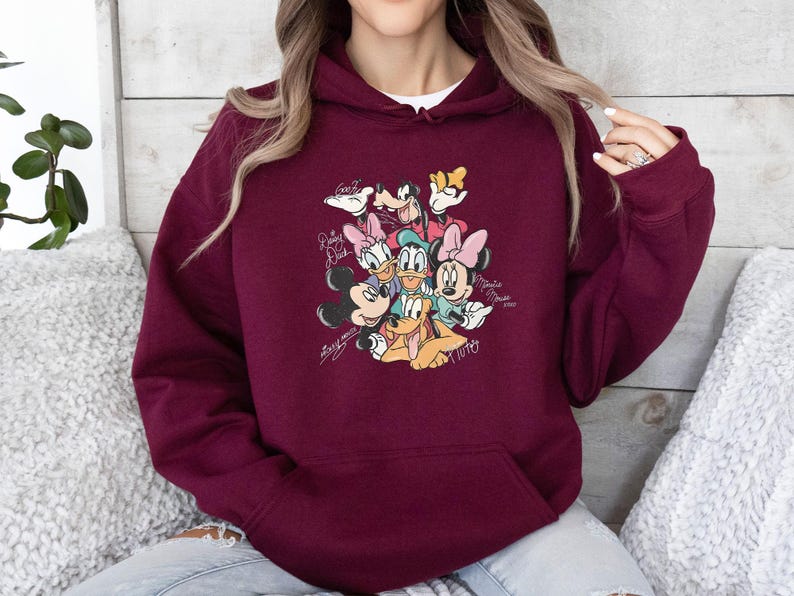 May include: Burgundy hooded sweatshirt featuring a colorful graphic of Disney characters including Mickey Mouse, Minnie Mouse, Donald Duck, Daisy Duck, Goofy, and Pluto. The design includes character signatures. A white undershirt is visible.