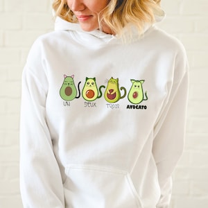 Può includere: Felpa con cappuccio bianca con una grafica di quattro gatti avocado. I gatti indossano orecchie da gatto e hanno noccioli di avocado per gli occhi. Il testo sotto i gatti dice "Un Deux Trois Avocado".