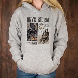Peut inclure: Un sweat &agrave; capuche gris avec un imprim&eacute; graphique noir et blanc d'un ch&acirc;teau, d'un livre et d'un dragon. Le texte "ONYX STORM" est imprim&eacute; en noir en haut du graphique. Le texte "BASGIATH WAR + COLLEGE" est imprim&eacute; en noir en bas du graphique.