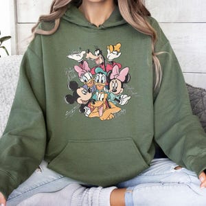 May include: Olive green hooded sweatshirt featuring a graphic of Disney characters including Mickey Mouse, Minnie Mouse, Donald Duck, Daisy Duck, Goofy, and Pluto. The design includes character signatures. The sweatshirt has a front pocket.