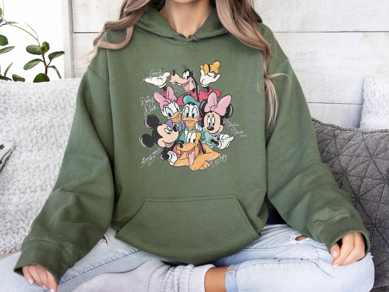 May include: Olive green hooded sweatshirt featuring a graphic of Disney characters including Mickey Mouse, Minnie Mouse, Donald Duck, Daisy Duck, Goofy, and Pluto. The design includes character signatures. The sweatshirt has a front pocket.