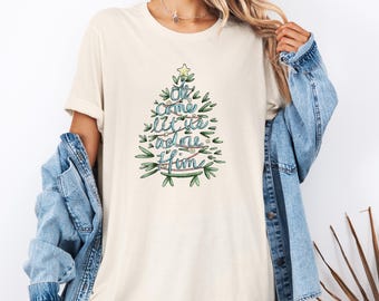 May include: Cream-colored t-shirt featuring a Christmas tree design with the words "Oh come let us adore Him" in a whimsical font. The tree is adorned with green leaves, blue and red accents, and a star on top. The shirt is a casual, comfortable style.