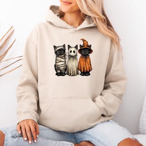 May include: Beige hoodie with a Halloween design of three black cats in costumes: a mummy, a ghost, and a witch. The cats are lined up, each in a different outfit. The hoodie has a drawstring hood and a front pocket.