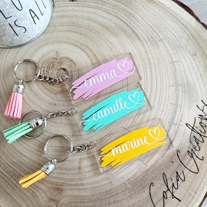 Puede incluir: Llaveros personalizados con los nombres Emma, Camille y Marine. Cada llavero presenta un diseño de pincelada colorida en rosa, turquesa y amarillo, con un corazón y un nombre en blanco. Los llaveros tienen anillos de metal plateado y borlas a juego.