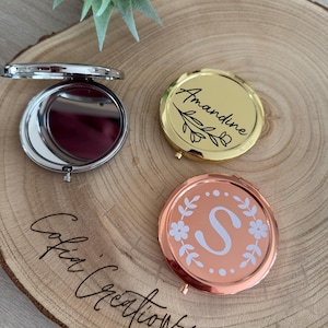 Peut inclure: Trois miroirs compacts personnalisés sur une surface en bois. L'un est ouvert, montrant le miroir, et les deux autres sont fermés. L'un est doré avec le nom "Amandine", et l'autre est or rose avec la lettre "S".