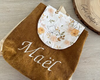 Op de afbeelding: Een bruine corduroy rugzak met een wit stoffen vak met een bloemenpatroon en een slapende vos. De rugzak heeft verstelbare banden en de naam "Maël" is geborduurd op de voorkant.