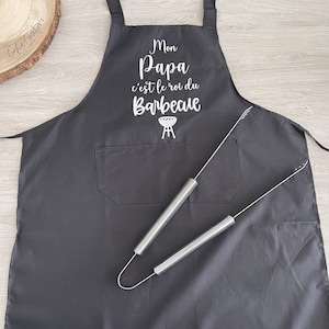 May include: Black apron with a white graphic that says "Mon Papa c'est le roi du Barbecue" with a grill icon. The apron has a pocket and two silver grilling tongs are on top of it.