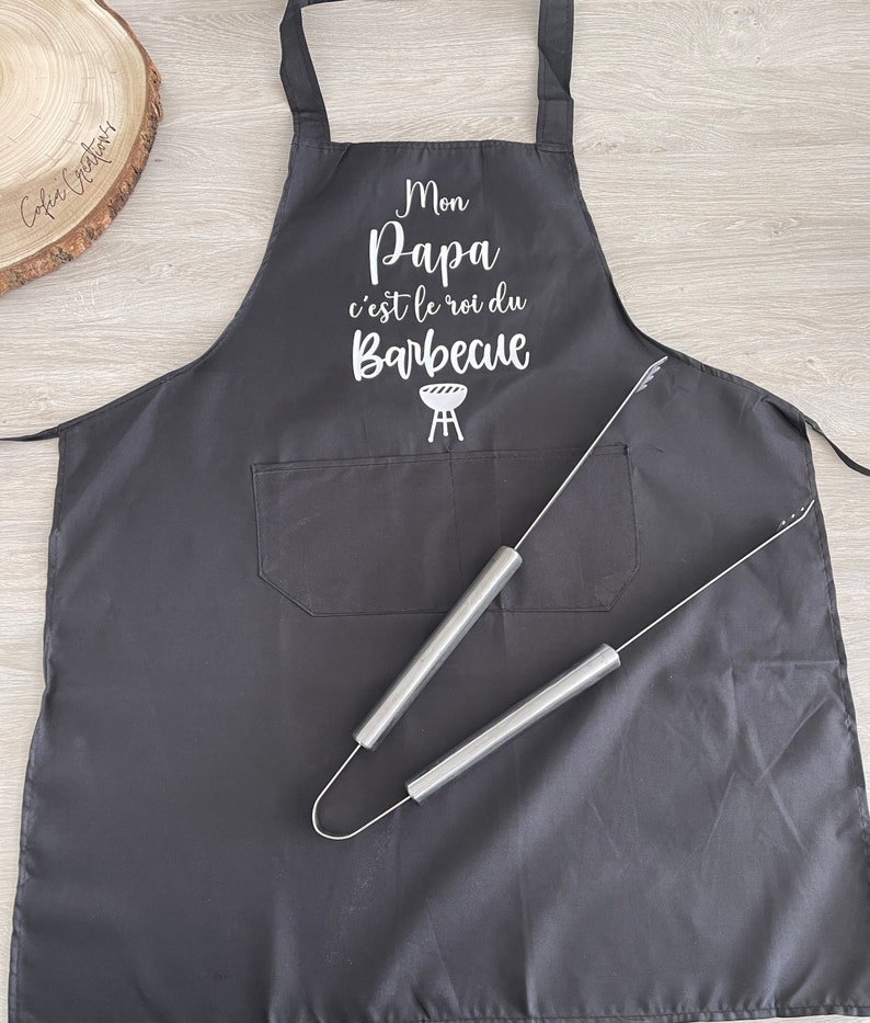 May include: Black apron with a white graphic that says "Mon Papa c'est le roi du Barbecue" with a grill icon. The apron has a pocket and two silver grilling tongs are on top of it.
