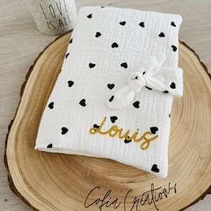 Può includere: Un bavaglino in mussola bianca con stampa a cuore nero e il nome "Louis" ricamato in glitter dorato. Il bavaglino è legato con un nastro in mussola bianca ed è esposto su una fetta di legno con il testo "Cofia Creations" inciso su di essa.
