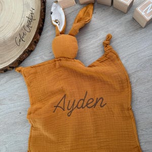 May include: An orange baby security blanket with a bunny head and ears, embroidered with the name "Ayden". Wooden letter blocks spell out "AYDEN". A wooden slice with the text "Cofia Creations" is also visible.