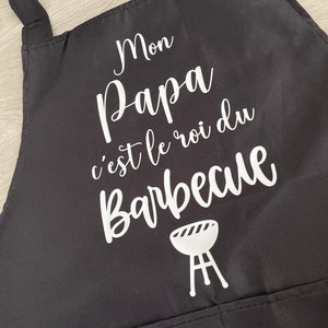 May include: Black apron with white text that reads "Mon Papa c'est le roi du Barbecue" and a white grill icon.