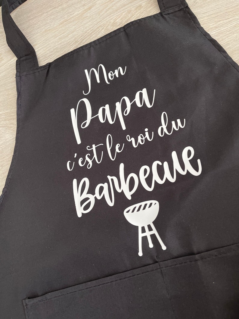 May include: Black apron with white text that reads "Mon Papa c'est le roi du Barbecue" and a white grill icon.