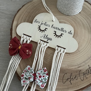 Op de afbeelding: Een houten wolkvormige haarbandhouder met de tekst "Les jolies barrettes de Alya" en een paar ogen. De houder heeft meerdere touwtjes die er vanaf hangen, en er hangen drie haarbandjes aan de touwtjes. De haarbandjes zijn rood en wit met bloemenpatronen.