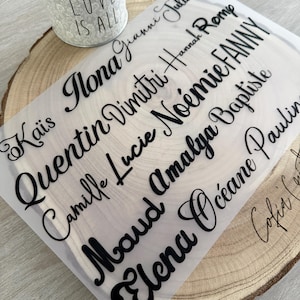 May include: A sheet of clear material with black cursive names, including Ilona, Quentin, and Elena, is displayed on a wooden slice. A small metal bucket with the words "LOVE IS ALL" is in the background.