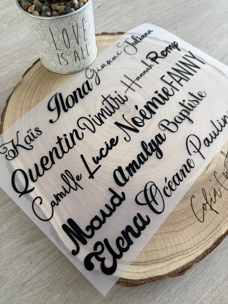 May include: A sheet of clear material with black cursive names, including Ilona, Quentin, and Elena, is displayed on a wooden slice. A small metal bucket with the words "LOVE IS ALL" is in the background.