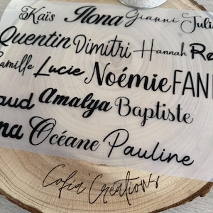May include: A clear sheet with multiple names in black script, including "Ka&iuml;s", "Mona", and "Quentin", is placed on a wooden slice. The words "Cofia Creations" are written in script at the bottom. A white candle holder with the words "LOVE IS ALL" is in the background.