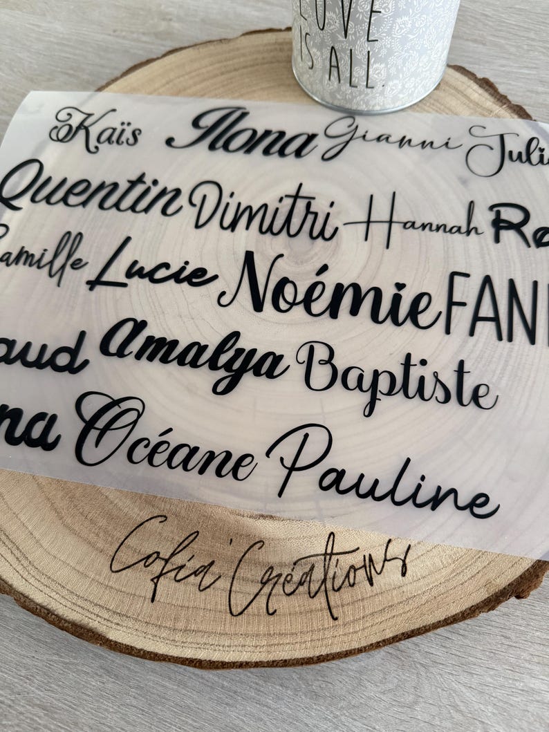 May include: A clear sheet with multiple names in black script, including "Ka&iuml;s", "Mona", and "Quentin", is placed on a wooden slice. The words "Cofia Creations" are written in script at the bottom. A white candle holder with the words "LOVE IS ALL" is in the background.