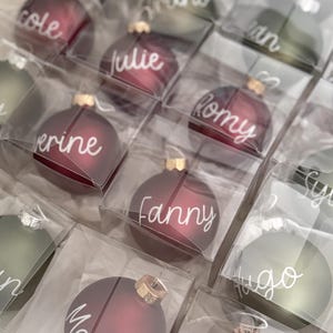 K&ouml;nnte beinhalten: Eine Sammlung runder, mattierter Weihnachtskugeln in Dunkelrot und Olivgr&uuml;n, einzeln verpackt in quadratischen Klarsichtschachteln. Jede Kugel ist mit einem wei&szlig;en, handgeschriebenen Namen versehen, darunter Cole, Julie, Romy, Fanny und Marie.