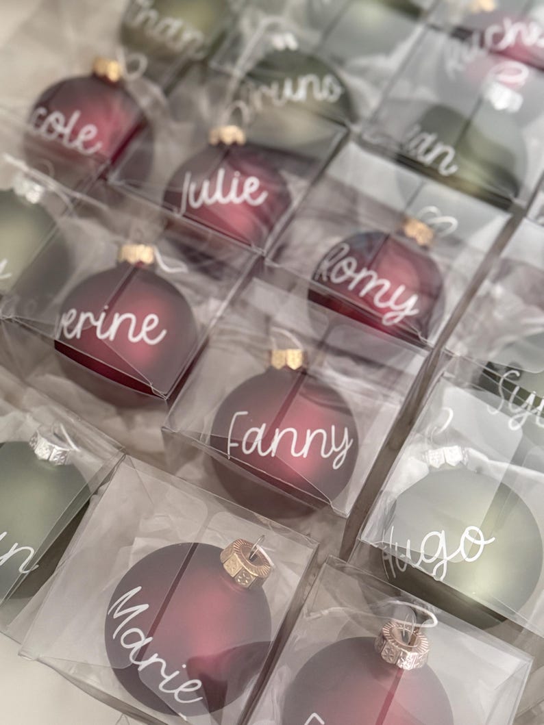 K&ouml;nnte beinhalten: Eine Sammlung runder, mattierter Weihnachtskugeln in Dunkelrot und Olivgr&uuml;n, einzeln verpackt in quadratischen Klarsichtschachteln. Jede Kugel ist mit einem wei&szlig;en, handgeschriebenen Namen versehen, darunter Cole, Julie, Romy, Fanny und Marie.