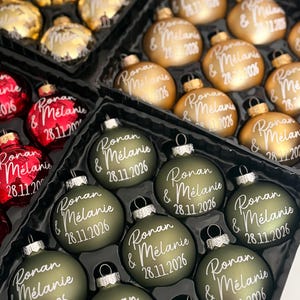 K&ouml;nnte beinhalten: Eine Sammlung runder Weihnachtskugeln in mattem Olivgr&uuml;n, Metallic-Gold und gl&auml;nzendem Rot. Jede Kugel ist mit dem wei&szlig;en Schreibschrift-Text "Ronan & M&eacute;lanie" und dem Datum "28.11.2026" auf der Vorderseite bedruckt.